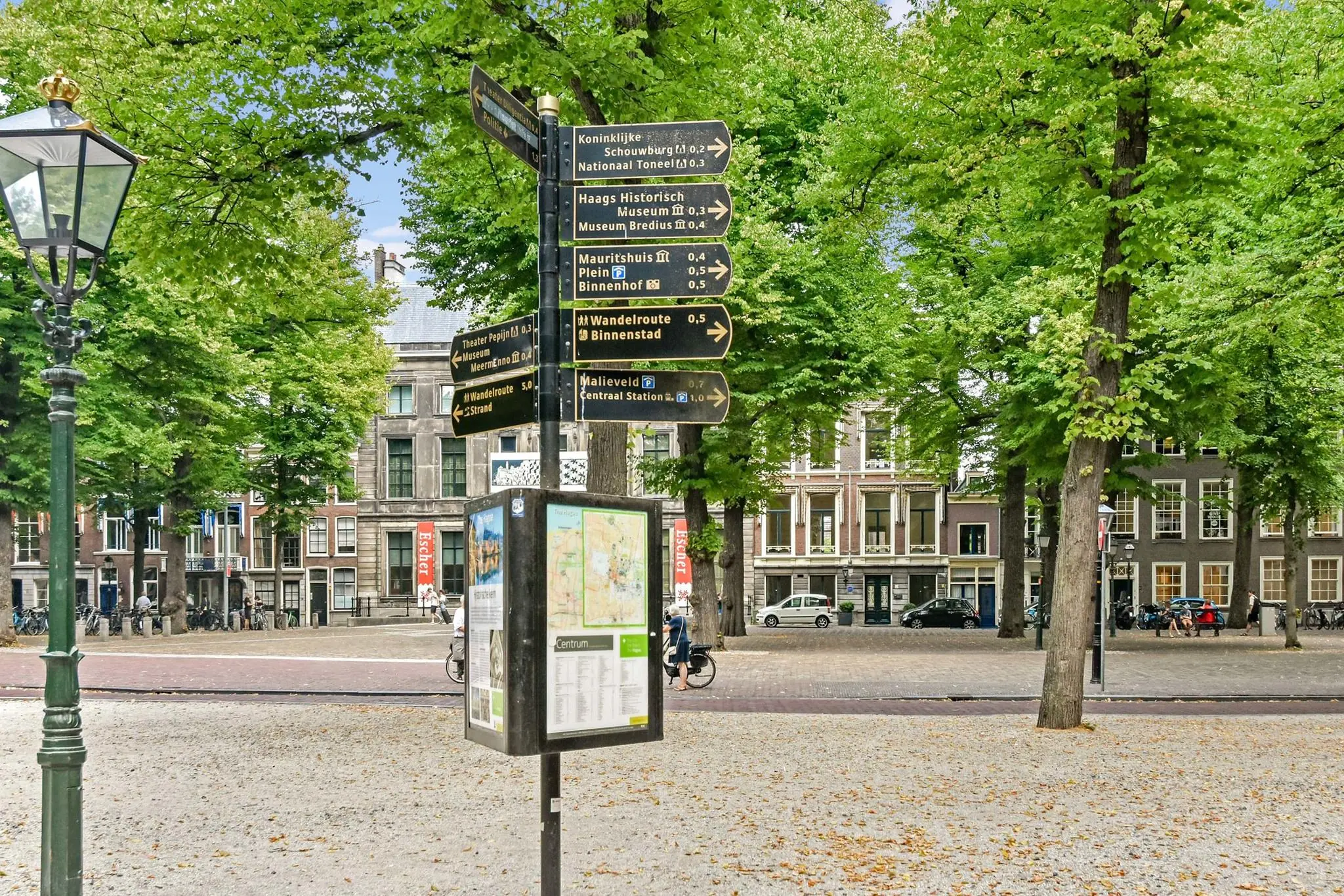 Wegwijzer en informatiebord op het Lange Voorhout in Den Haag, omgeven door bomen en historische panden.
